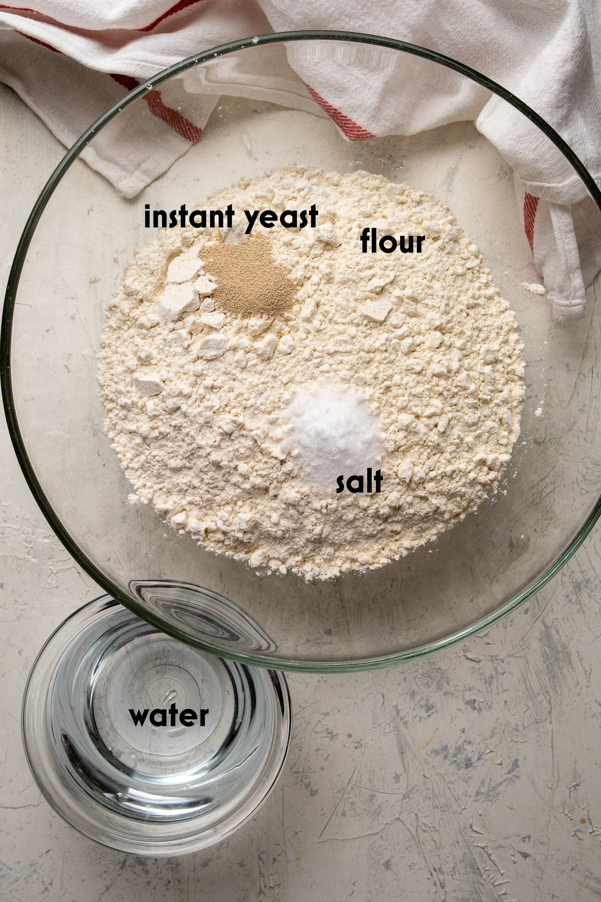 Flour, instant yeast and salt in a large glass bowl and water in a smaller bowl on the side on a light background.