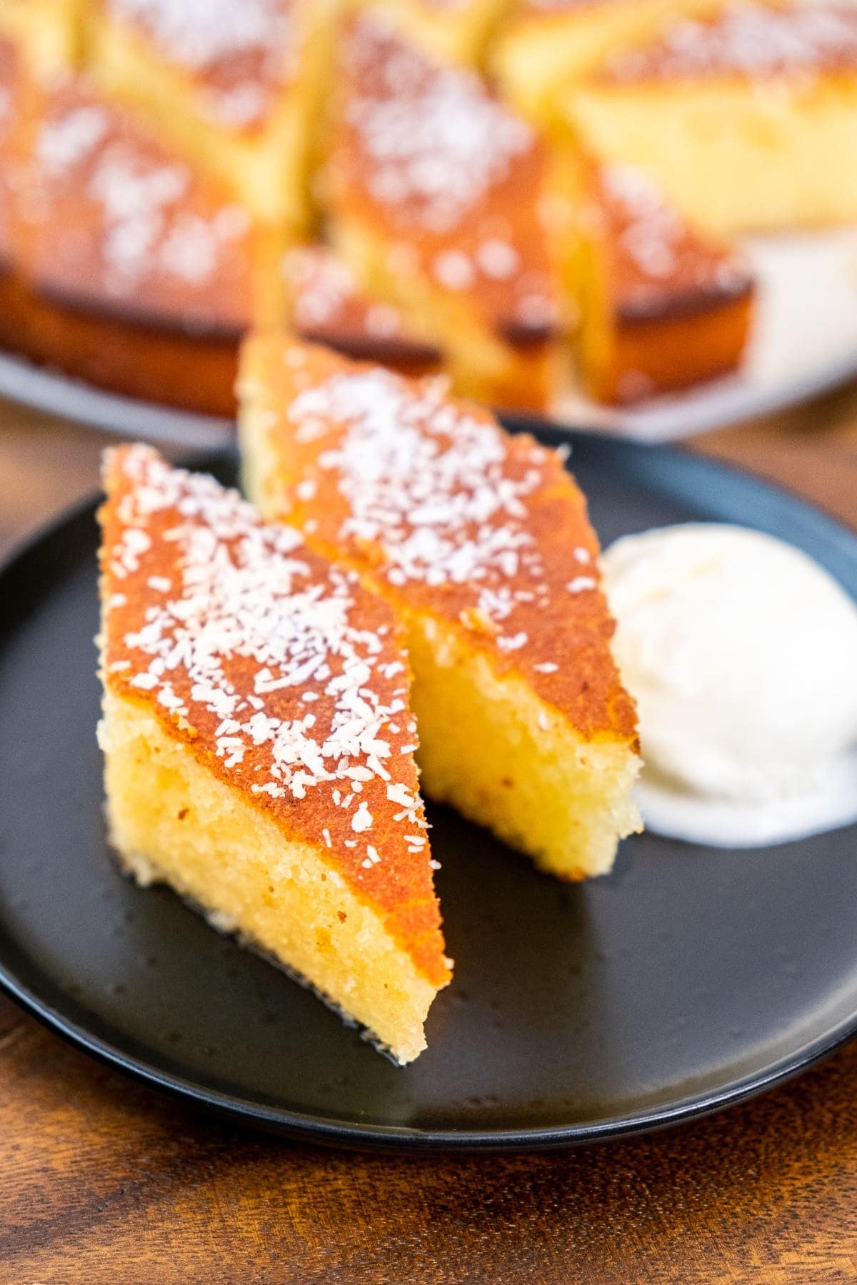 Two slices of Turkish revani cake garnished with desiccated coconut on a black plate and ice cream on the side.