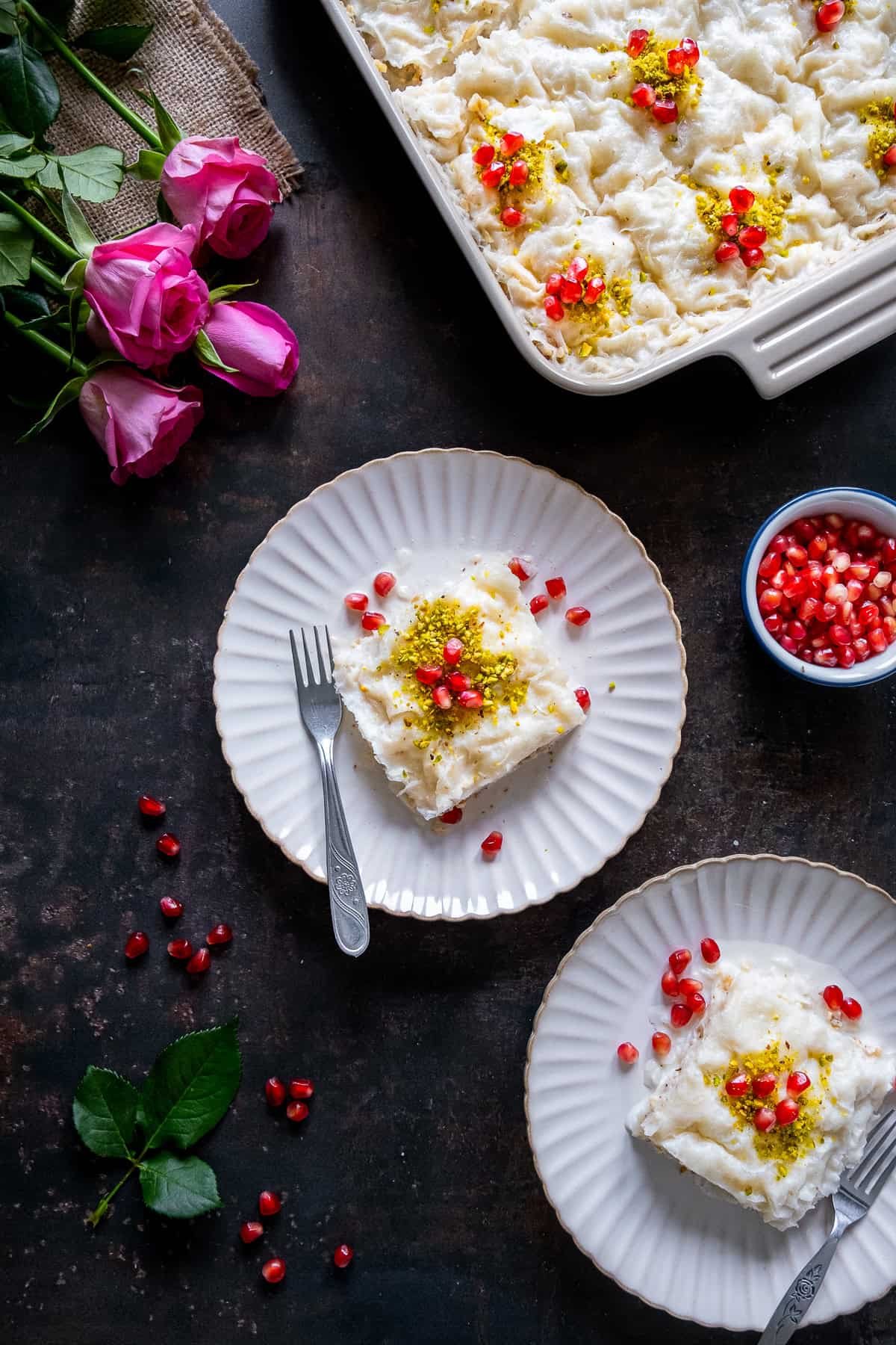 Two plates of Turkish Güllaç dessert served on white scalloped plates, garnished with ground pistachios and pomegranate seeds, with a baking dish of the dessert and pink roses in the background.