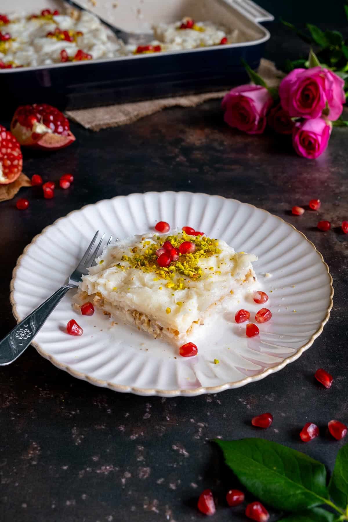 A close-up of a slice of Turkish Güllaç dessert on a white plate, topped with crushed pistachios and pomegranate seeds, with a baking dish and pink roses in the background.