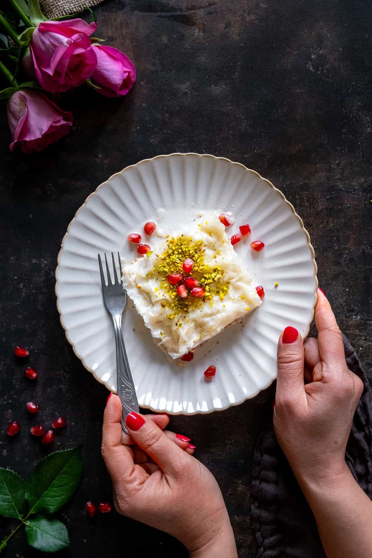 A person holding a plate with a slice of Turkish Güllaç dessert, topped with crushed pistachios and pomegranate seeds, with pink roses and scattered pomegranate arils on a dark surface.