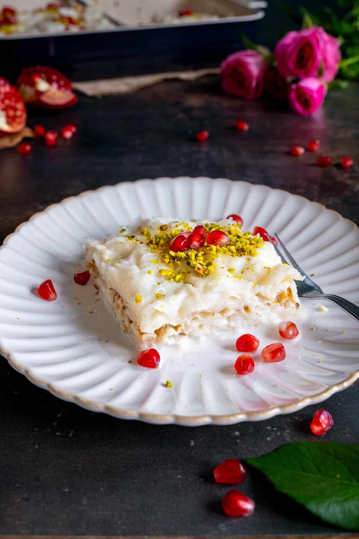 A close-up of a creamy slice of Turkish Güllaç dessert on a white plate, topped with ground pistachios and pomegranate seeds, with pink roses and pomegranate pieces in the background.
