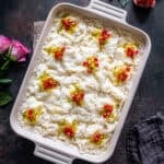 A large rectangular dish of Turkish Güllaç dessert, cut into squares and decorated with crushed pistachios and pomegranate seeds, with pink roses and a dark cloth nearby.