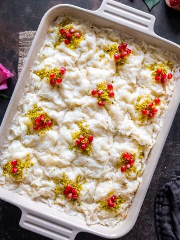 A large rectangular dish of Turkish Güllaç dessert, cut into squares and decorated with crushed pistachios and pomegranate seeds, with pink roses and a dark cloth nearby.