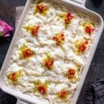 A large rectangular dish of Turkish Güllaç dessert, cut into squares and decorated with crushed pistachios and pomegranate seeds, with pink roses and a dark cloth nearby.