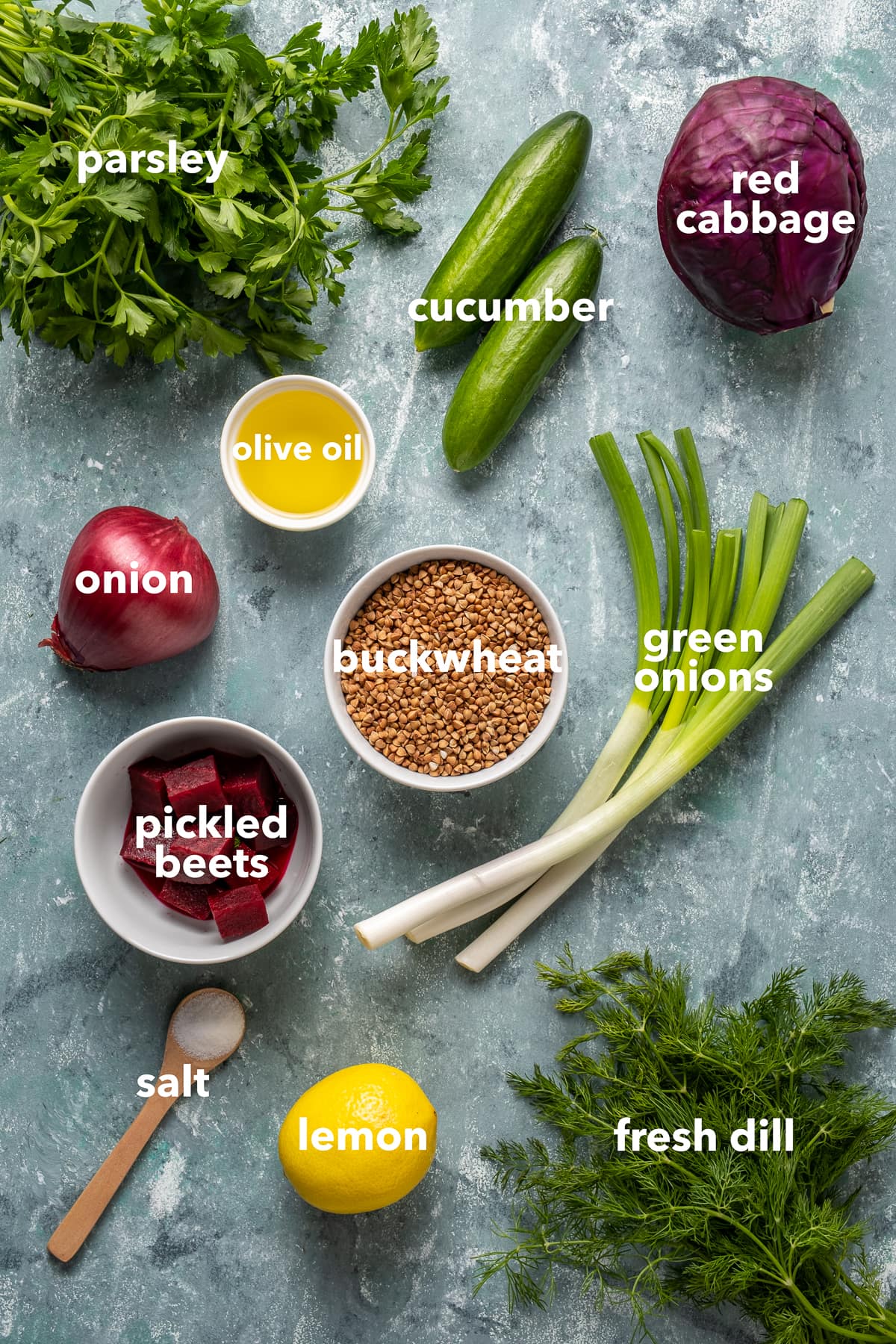 Buckwheat groats, pickled beets, red onion, a bunch of parsley, a bunch of fresh dill, baby cucumbers, red cabbage, olive oil, green onions, lemon and salt in a wooden teaspoon on a light background.