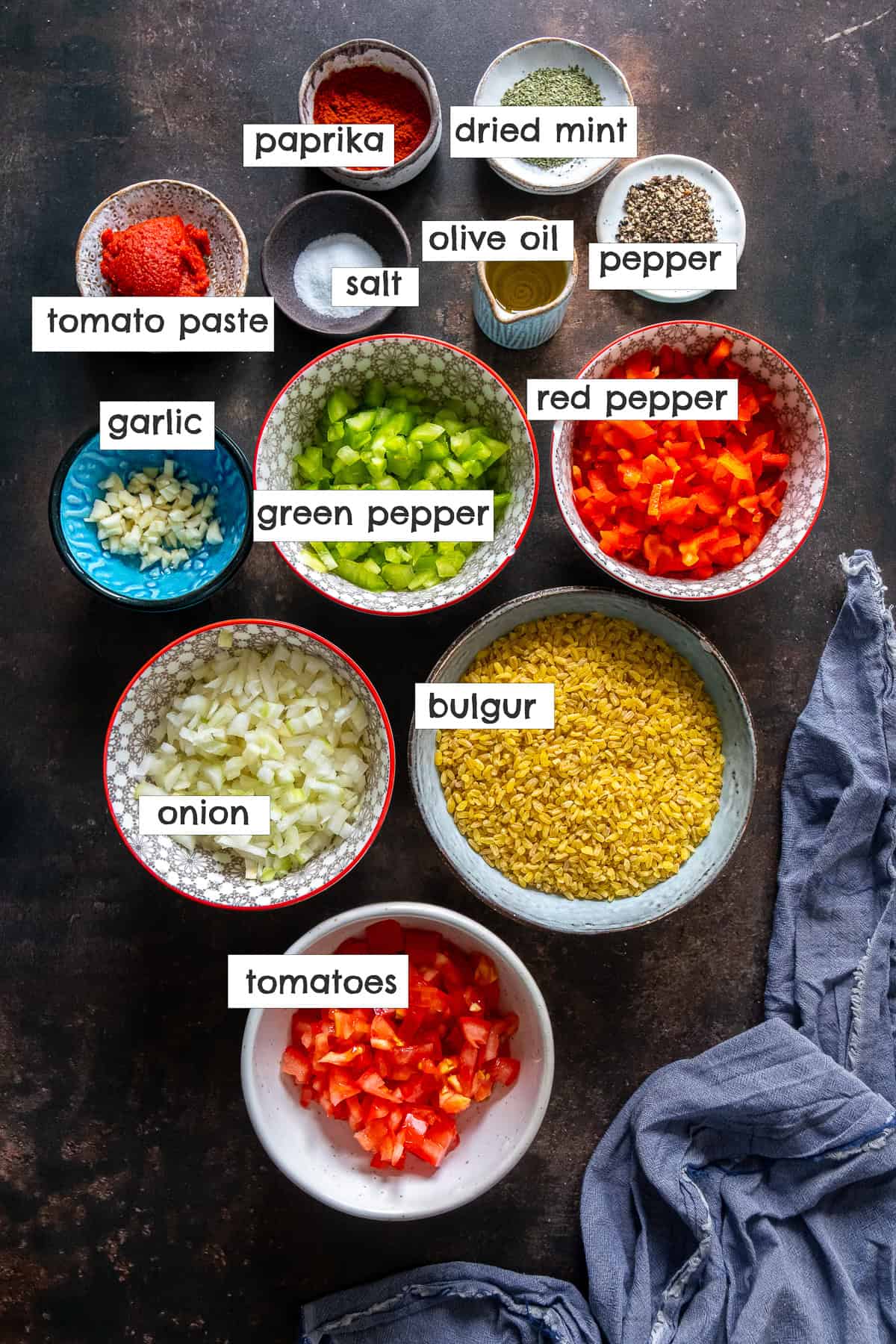 Labeled ingredients for Turkish bulgur pilaf, including bulgur, onion, garlic, green pepper, red pepper, tomatoes, tomato paste, paprika, dried mint, olive oil, salt, and pepper, arranged in bowls on a dark surface.