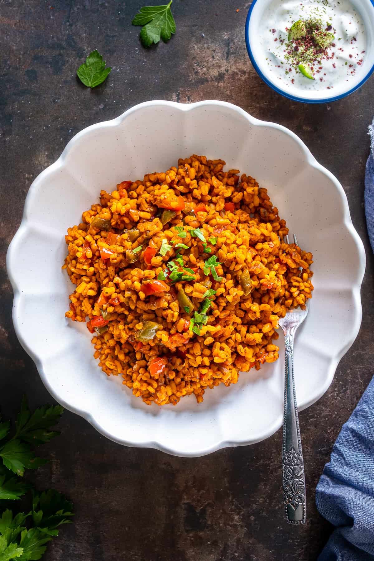 A white plate of Turkish bulgur pilaf with peppers and parsley garnish, served with a fork, alongside a small bowl of cacik on a dark background.
