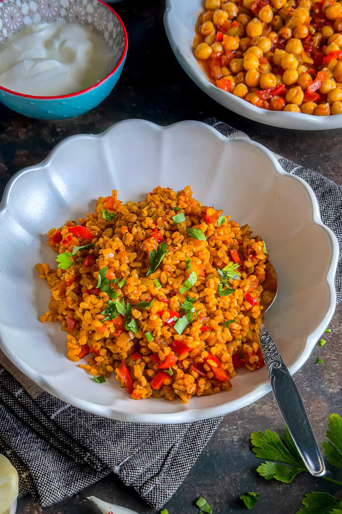 A bowl of Turkish bulgur pilaf with red peppers and parsley, served with a spoon, alongside dishes of yogurt and chickpea stew.