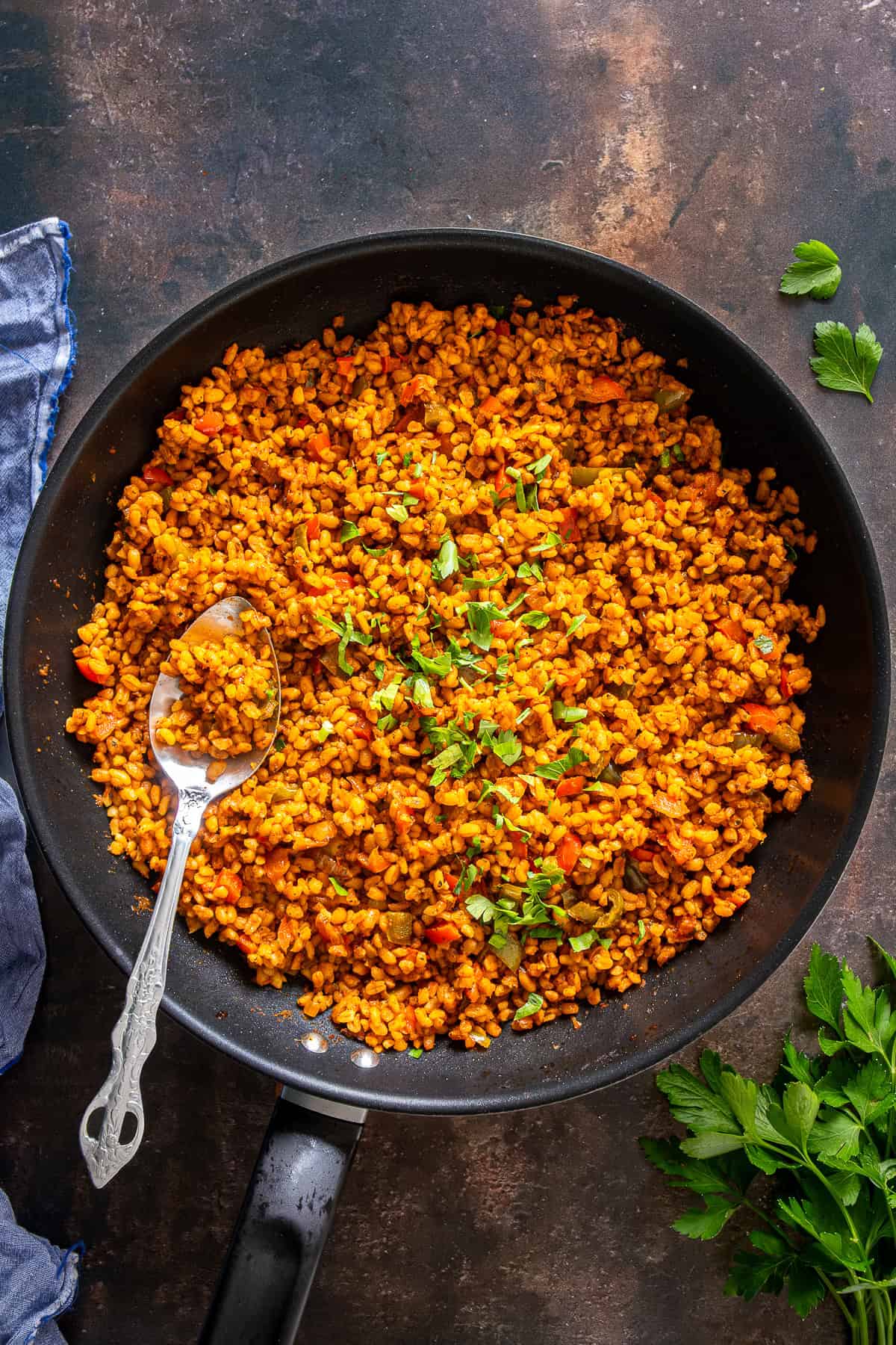 A pan of freshly cooked Turkish bulgur pilaf with peppers, garnished with parsley, and served with a spoon.