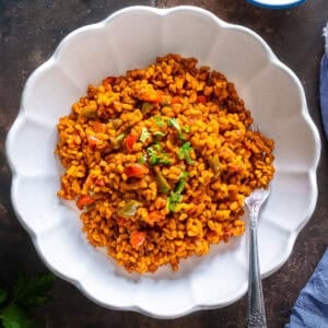 A plate of Turkish bulgur pilaf with red and green peppers, garnished with parsley, served in a white scalloped dish with a fork.