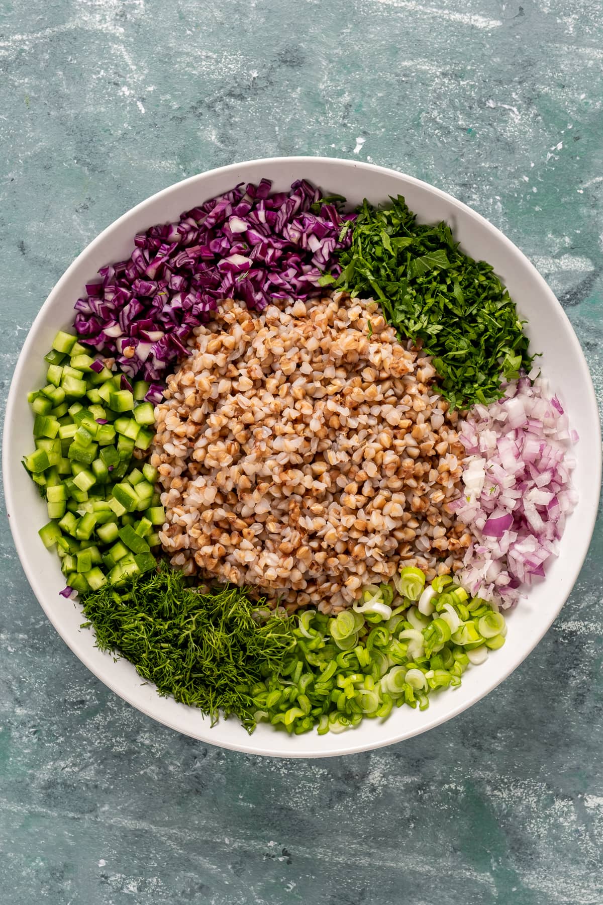 Cooked buckwheat, finely chopped red onion, red cabbage, parsley, fresh dill, green onions and cucumbers all in a white salad bowl.