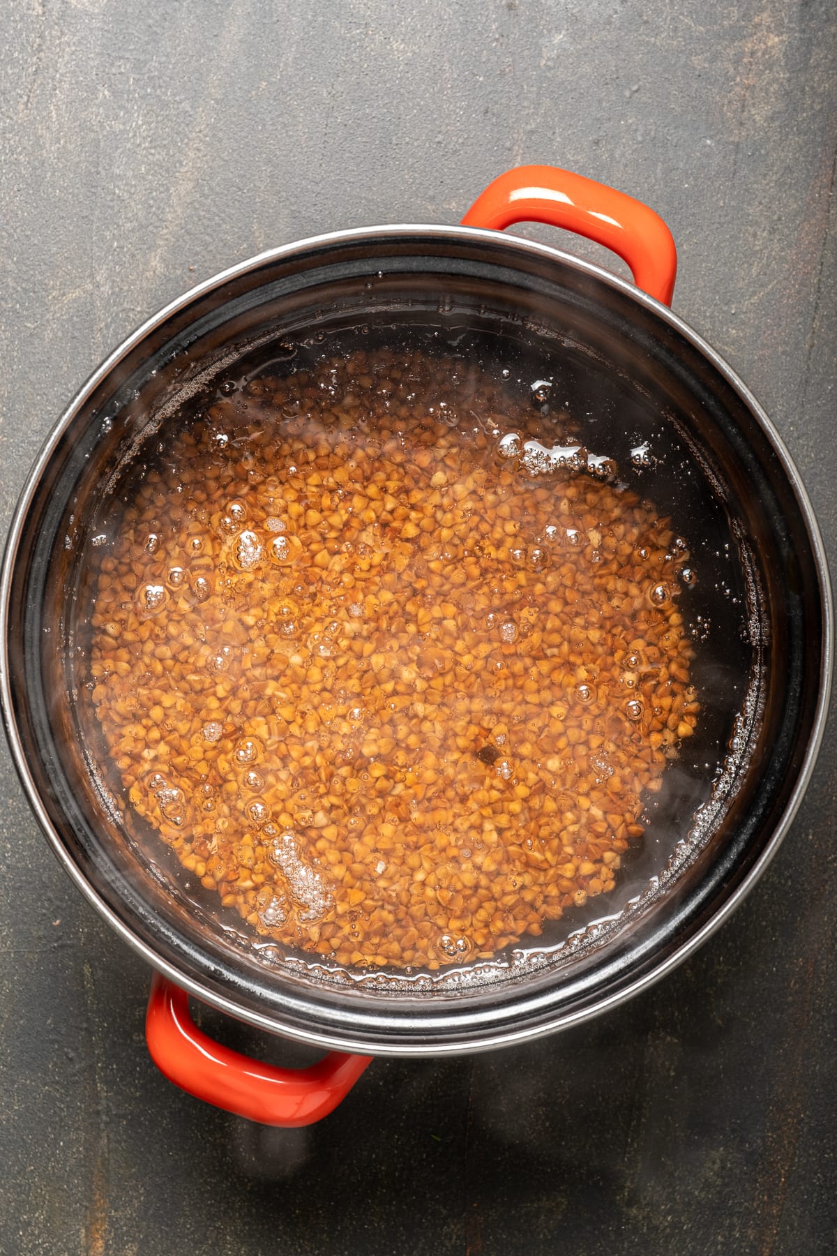 Buckwheat groats and water in a black pan with red handles.