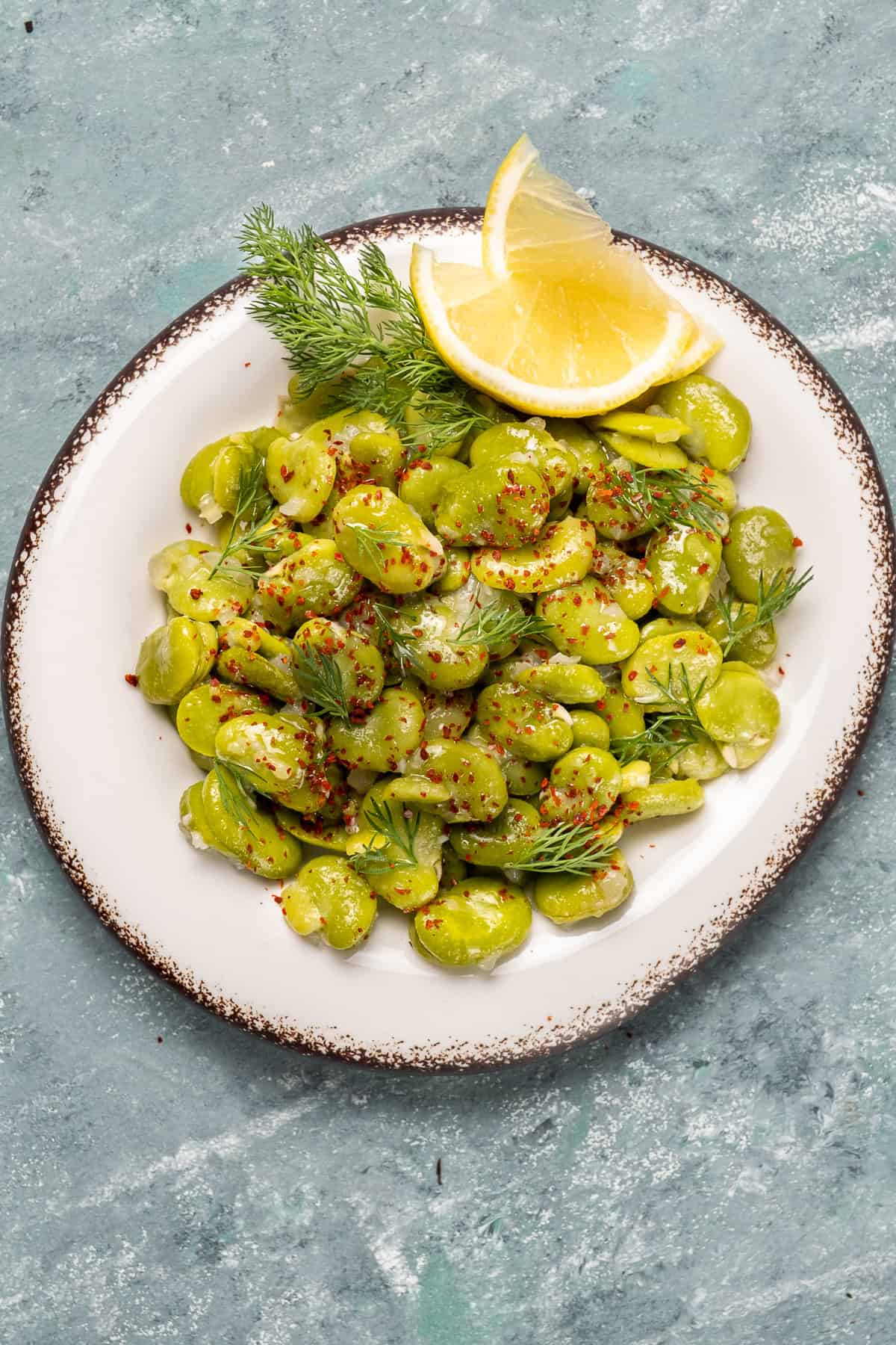 Plate of fresh fava beans in olive oil garnished with lemon wedges, dill, and red pepper flakes.