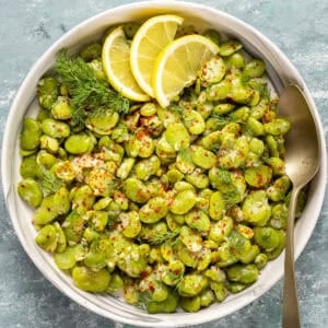 Bowl of fresh fava beans in olive oil topped with lemon slices, dill, and red pepper flakes.