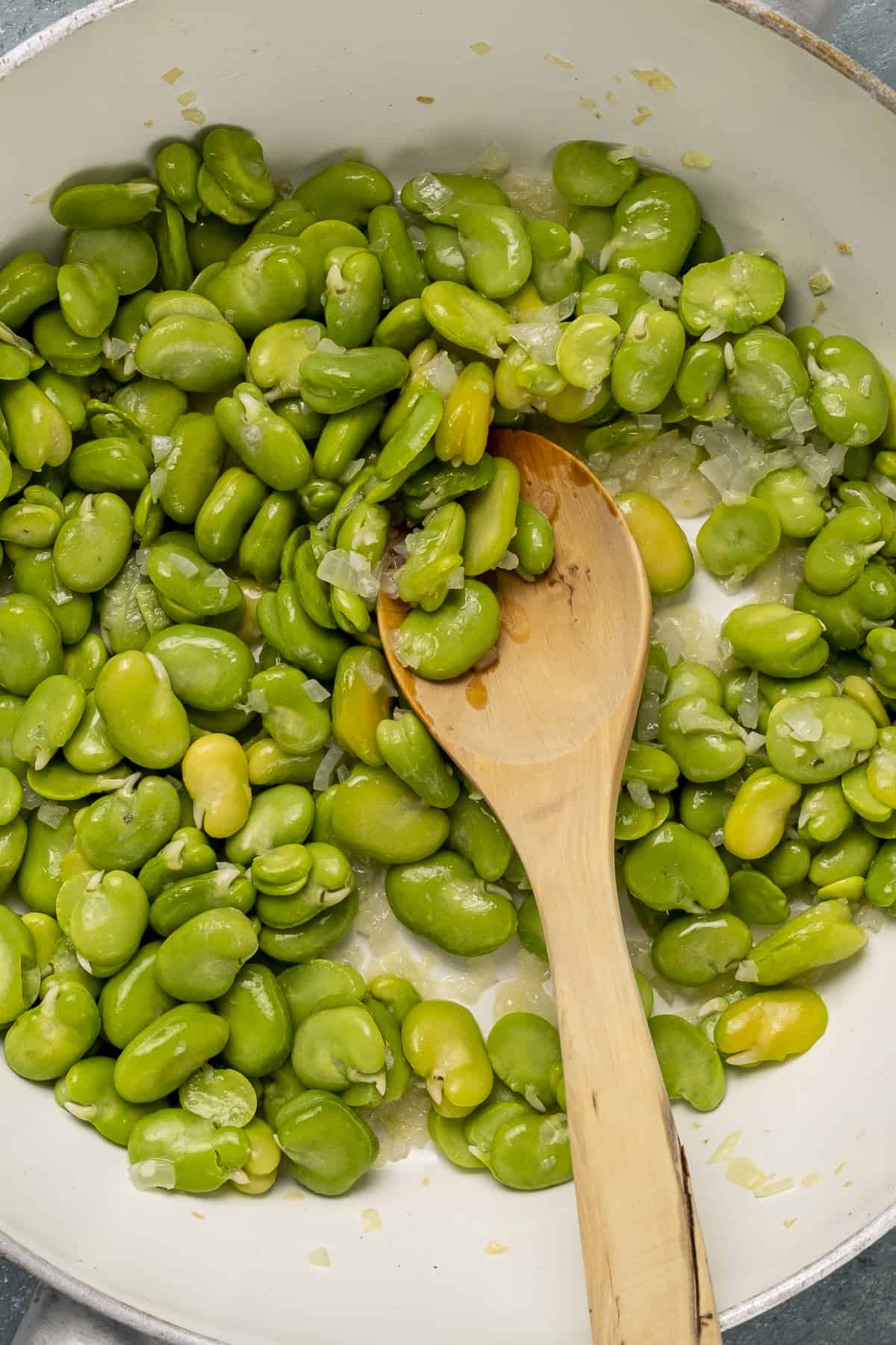 Green fresh fava beans cooking in a pan and a wooden spoon in it.