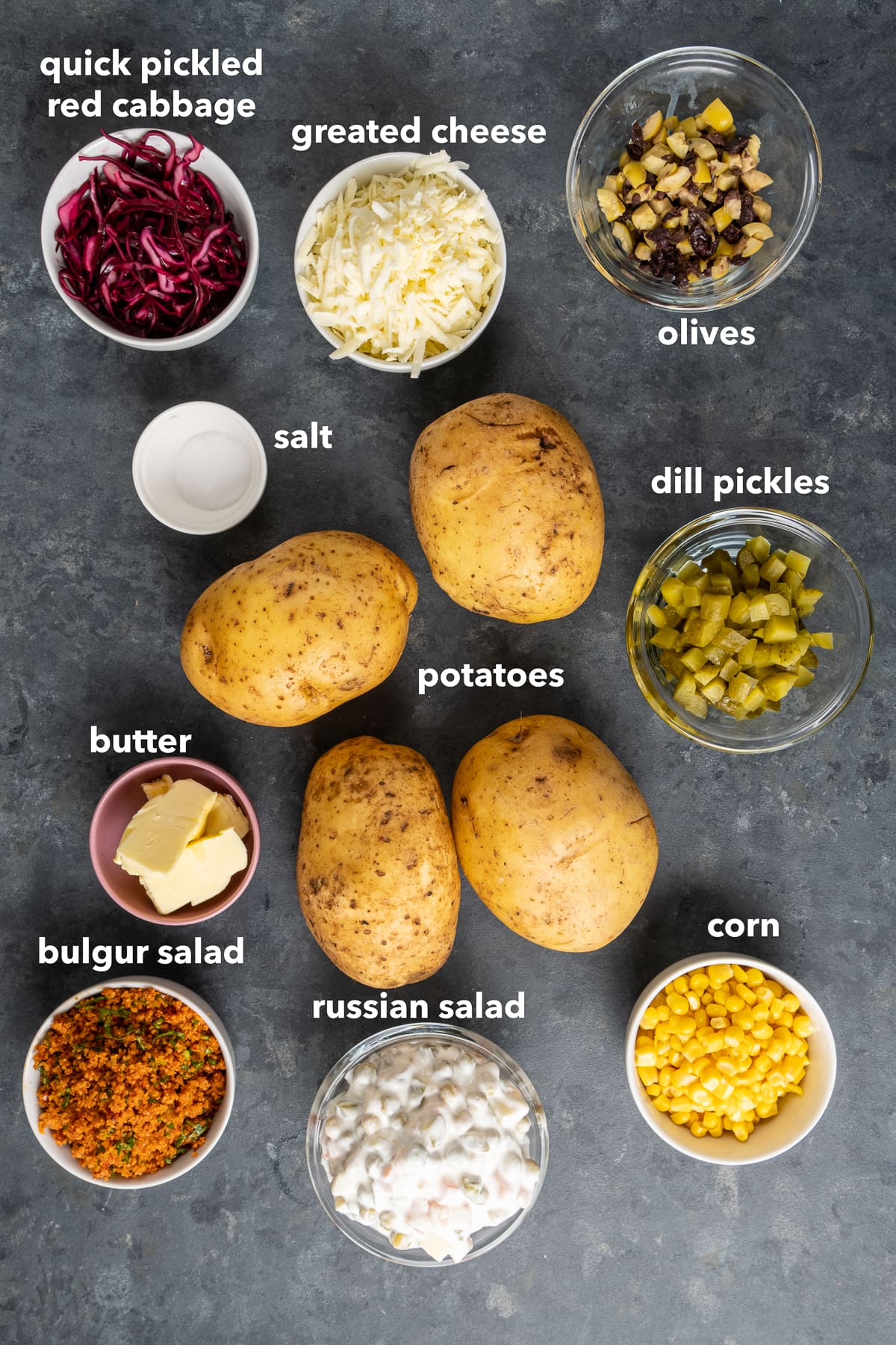 Potatoes, dill pickles, red cabbage, grated cheese, salt, chopped olives, Russian salad, bulgur salad and butter on a dark background.