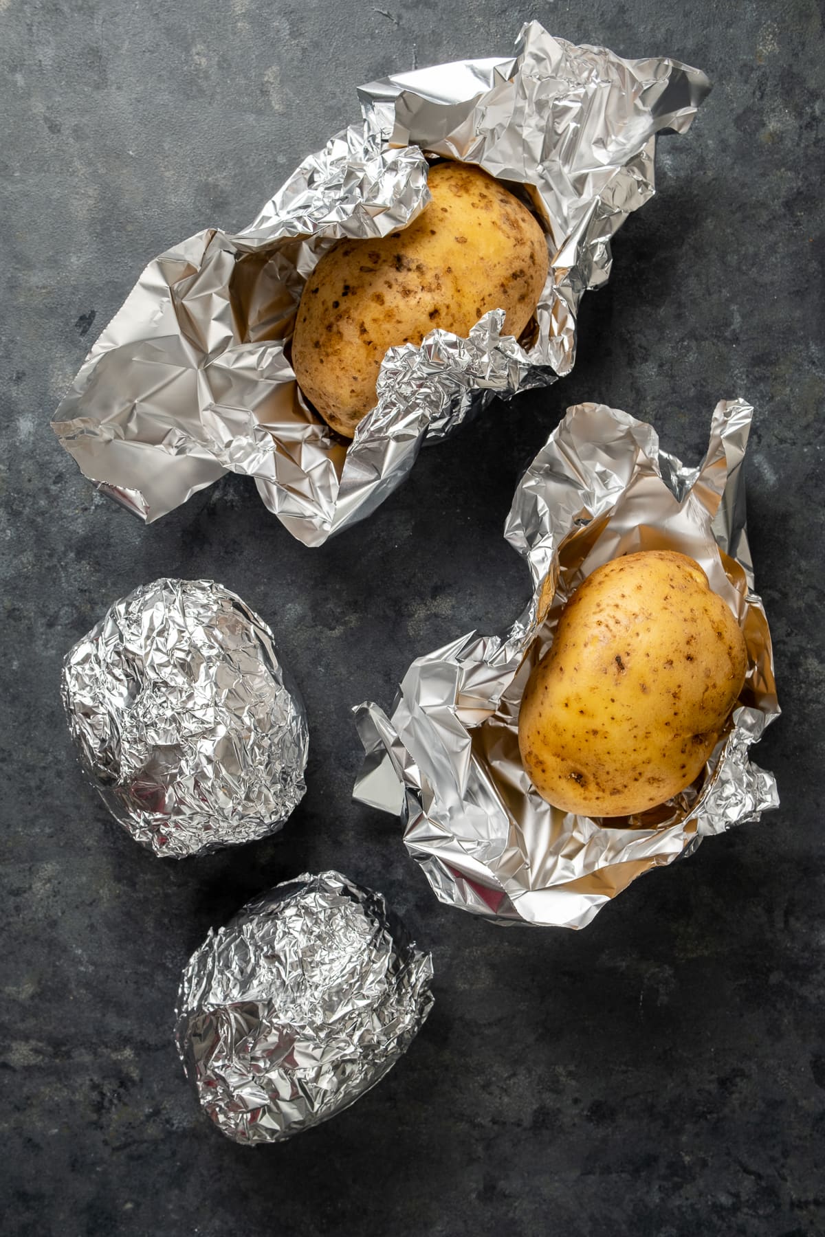Two potatoes wrapped in foil, two potatoes are half wrapped with foil on a dark background.