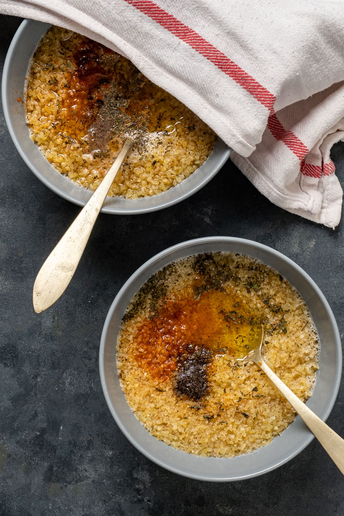 Medium bulgur soaked in water with spices in two bowls with spoons insde them. One of them is partly covered with a kitchen towel.