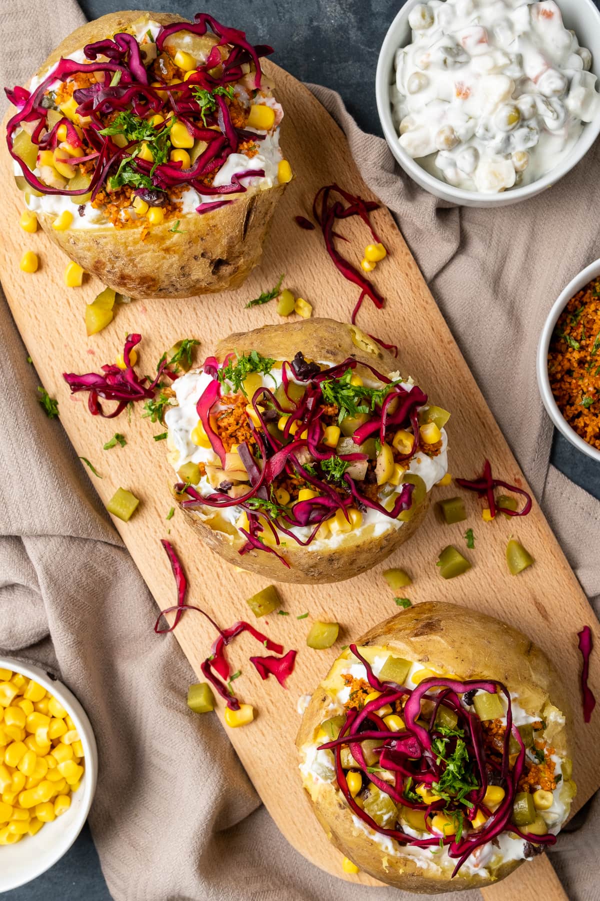 Loaded baked potatoes called kumpir on a wooden board. Russian salad, sweet corn and bulgur salad in separate bowls on the side.