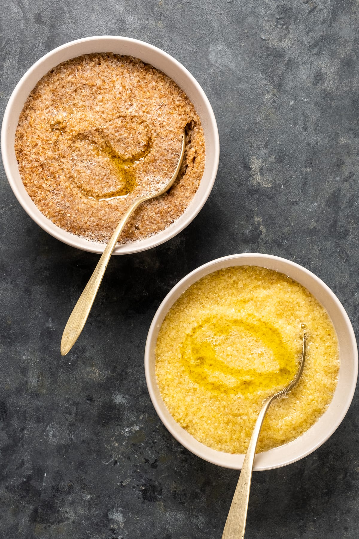 Red fine bulgur and white fine bulgur soaked in water and olive oil in two white bowls with spoons inside them.