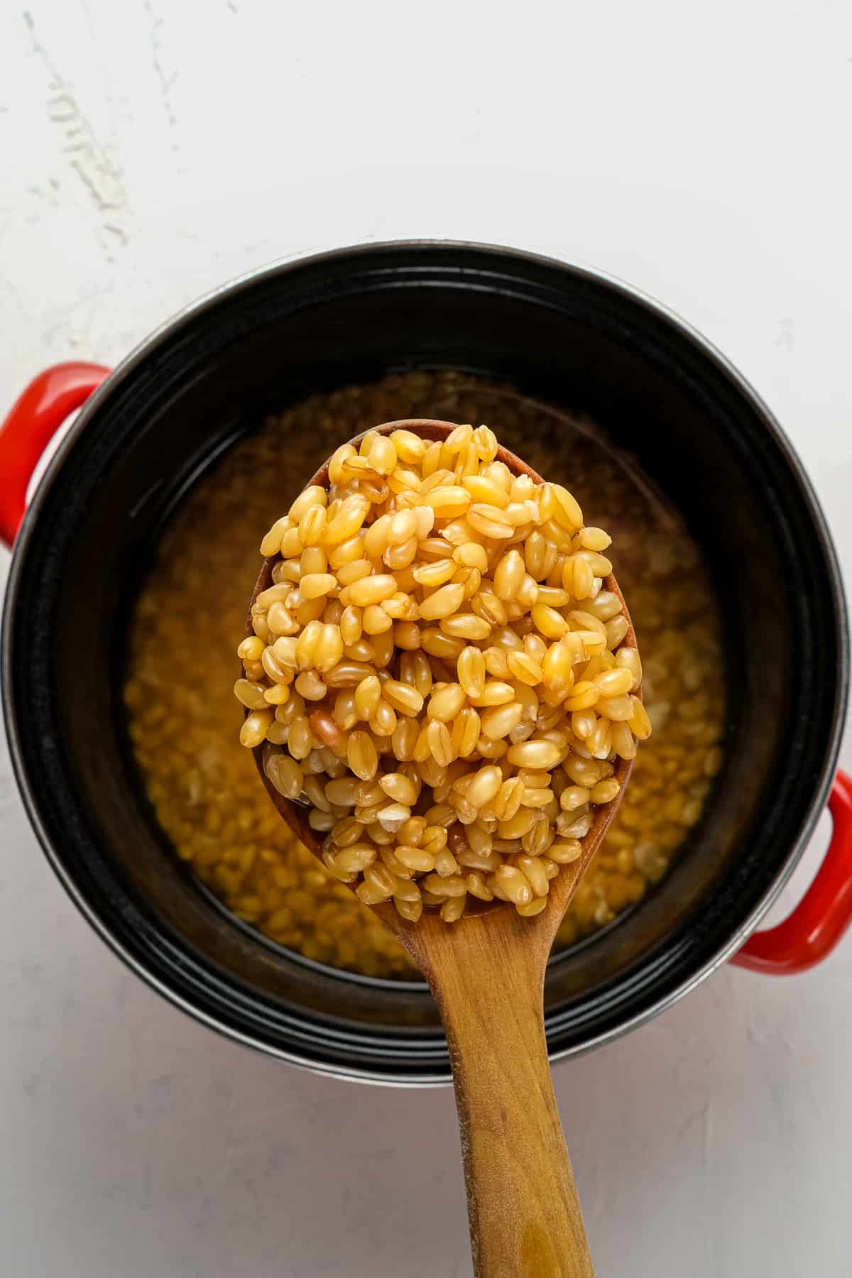 Cooked wheat berries in a wooden spoon over a black enamel pan with red handles.