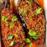 Two Turkish karniyarik eggplants filled with a savory ground beef and vegetable mixture, topped with parsley and served on a white plate with tomato sauce.