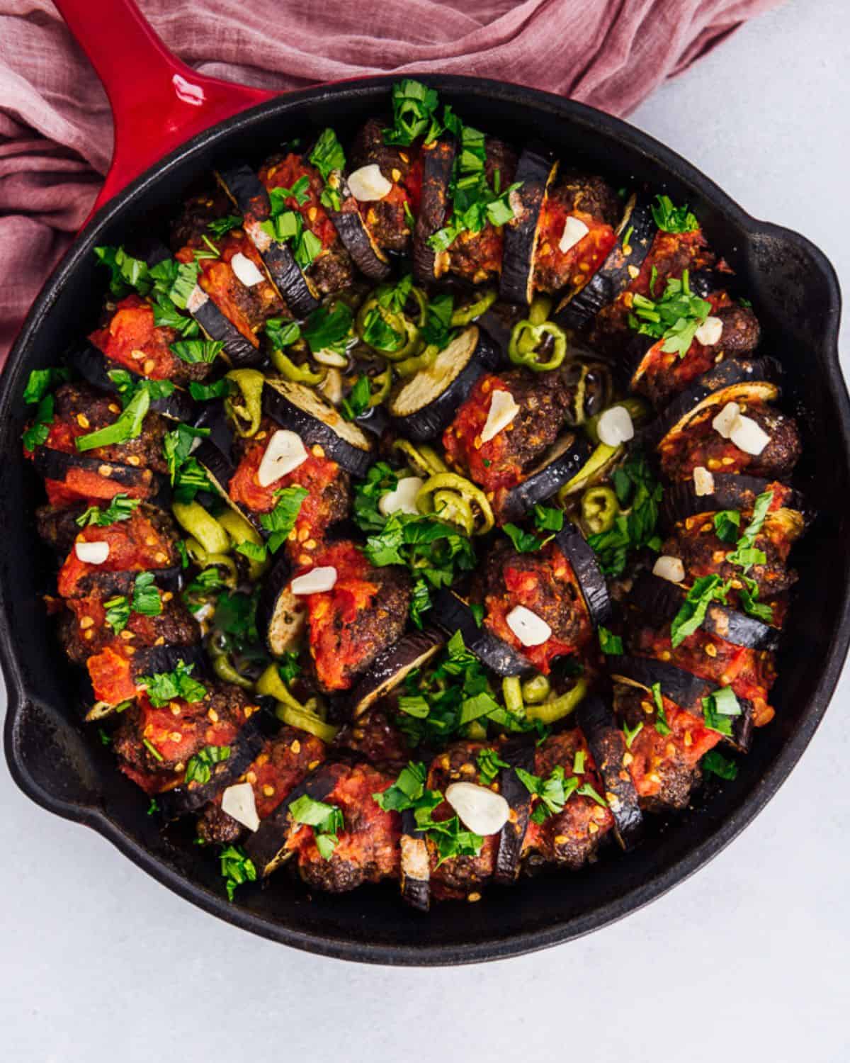 An image of eggplant kebab served in a black pan, topped with parsley, garlic, and green peppers.