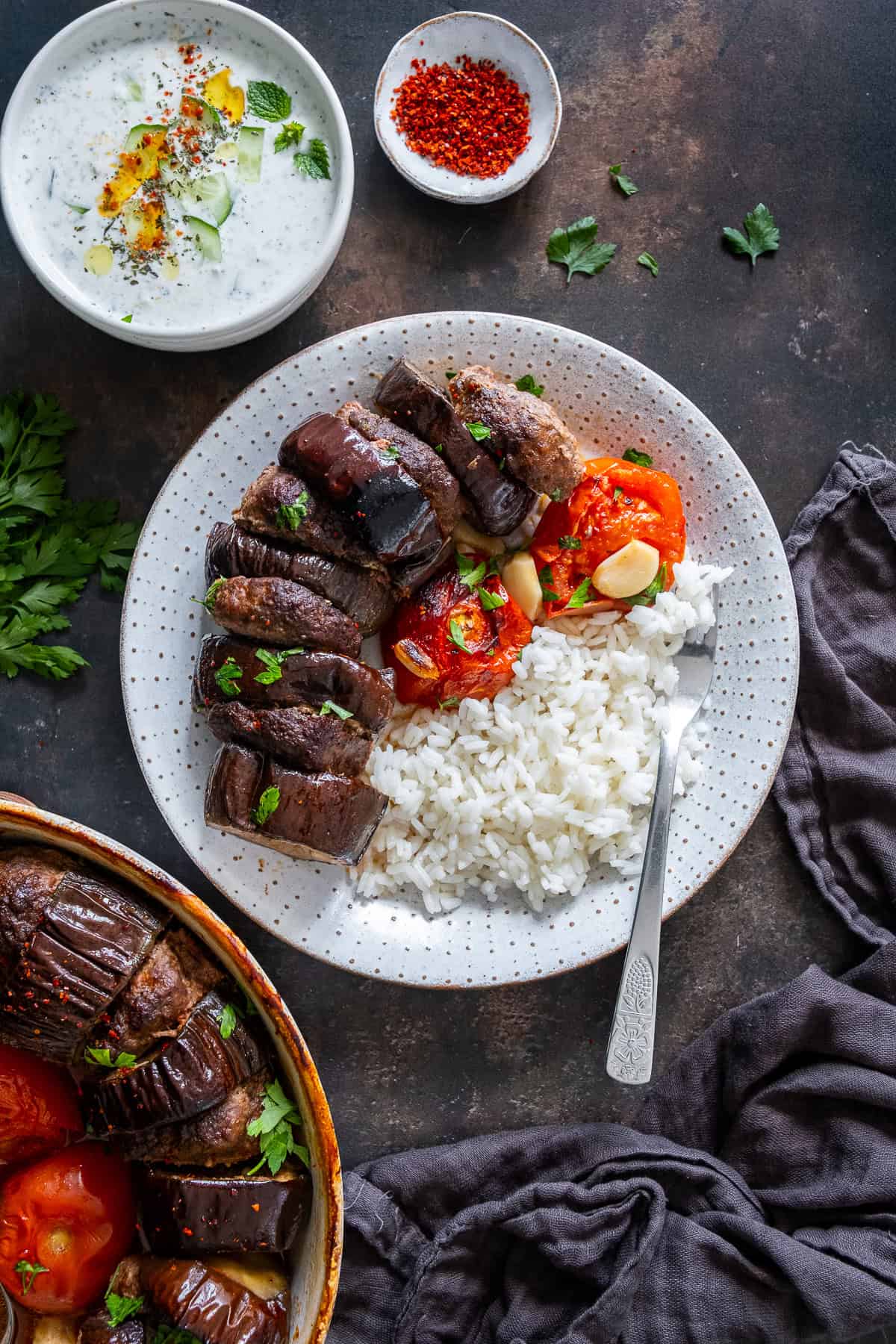 A serving of Turkish eggplant kebab with rice pilaf on a plate, with a bowl of cacık and the baking dish in the background.