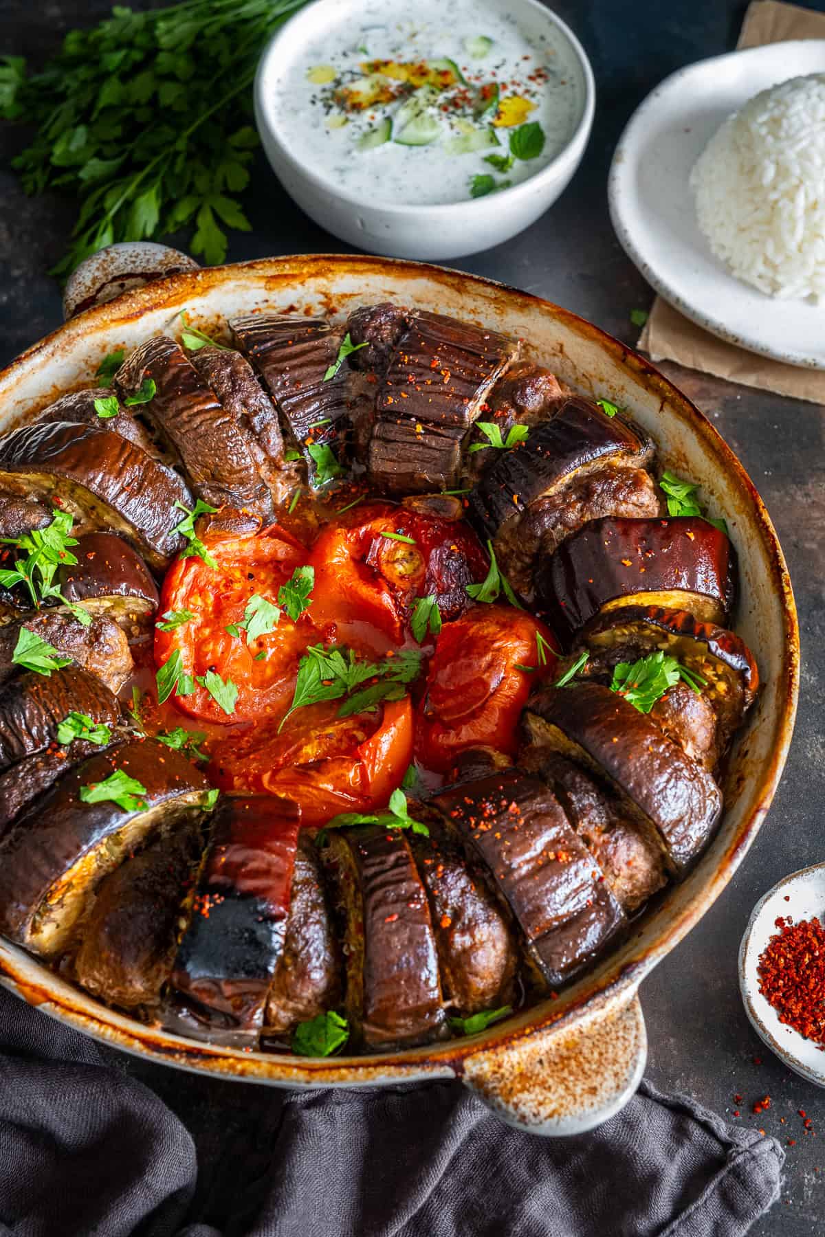 Close-up of Turkish eggplant kebab baked with meatballs, tomatoes and garlic in a round baking dish, served with rice pilaf and cacık in the background.