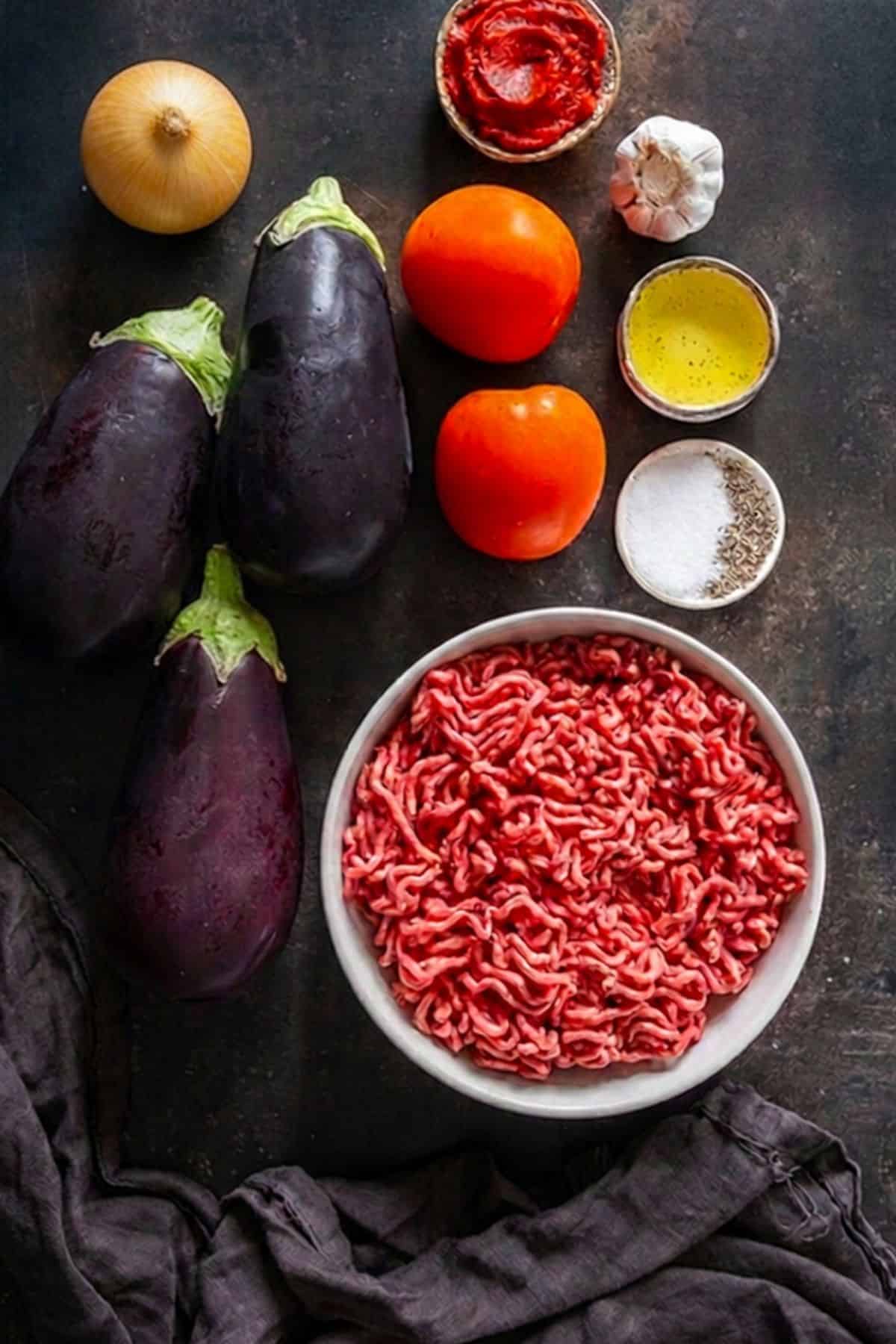 Ingredients for Turkish eggplant kebab arranged on a dark surface, including ground beef, eggplants, tomatoes, garlic, onion, tomato paste, olive oil, salt and black pepper.