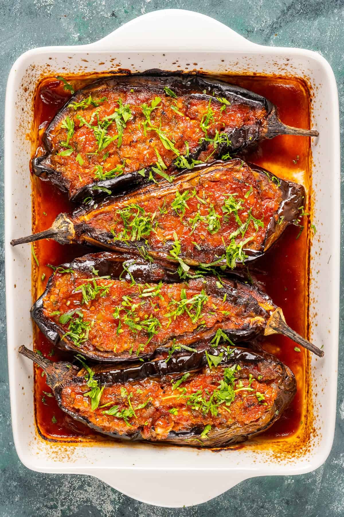Four baked stuffed eggplants in a white dish, topped with tomato sauce and chopped parsley, sitting in a rich olive oil and tomato base.