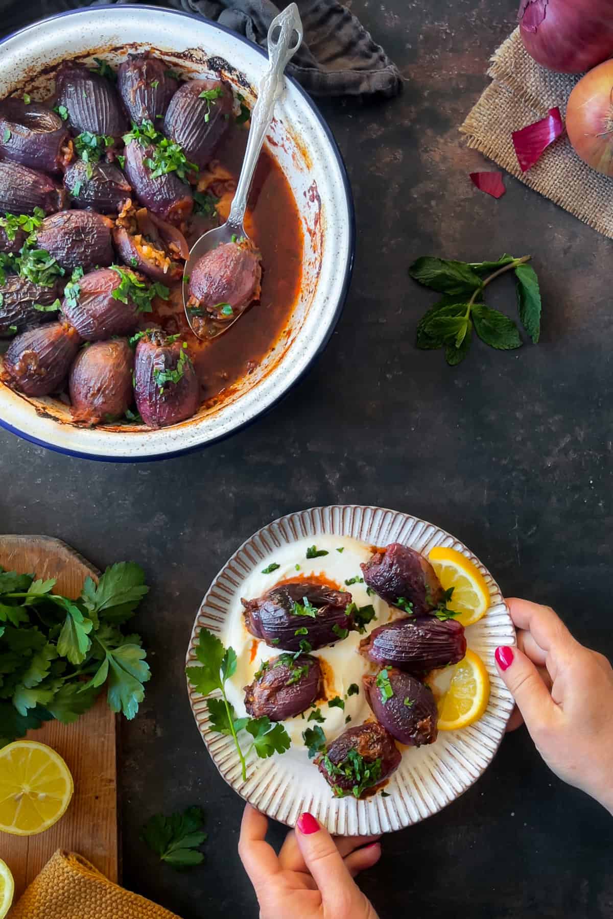 A serving of Sogan Dolmasi (Turkish Stuffed Onions) plated on yogurt, garnished with fresh parsley and lemon slices. The main dish is in the background with a spoon scooping one stuffed onion.