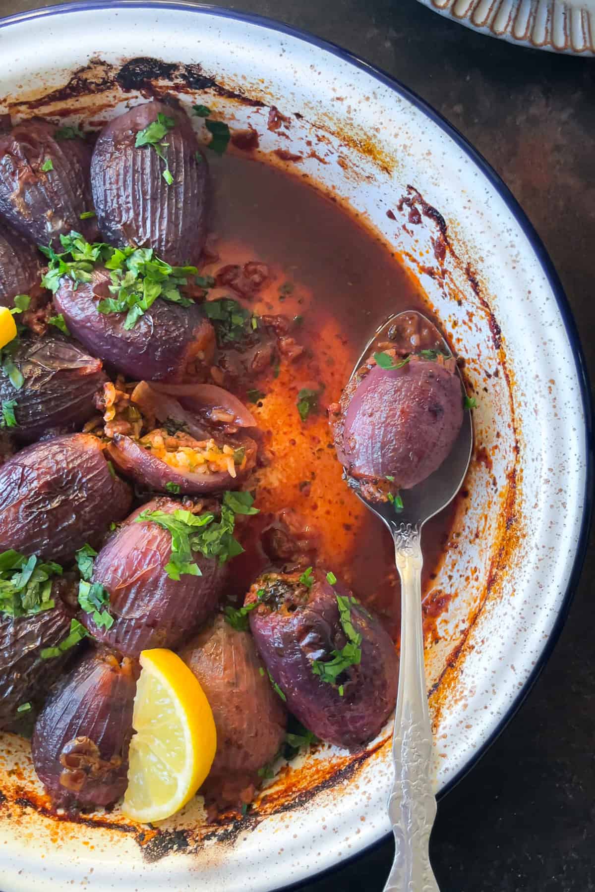 A close-up of Sogan Dolmasi (Turkish Stuffed Onions) in a baking dish, garnished with parsley and lemon. A spoon lifts one stuffed onion, showing the rich, flavorful sauce.