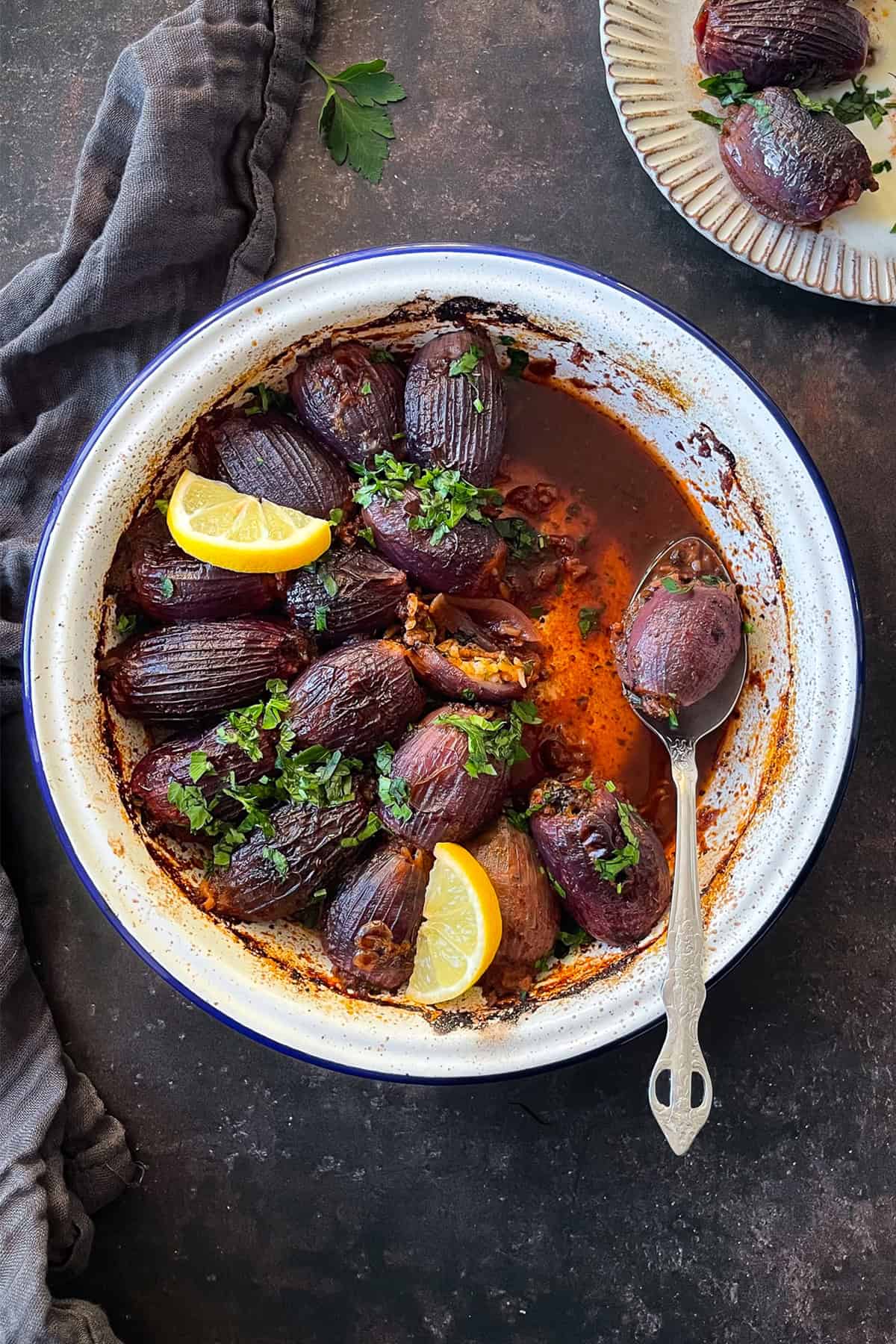A rustic enamel dish filled with Sogan Dolmasi (Turkish Stuffed Onions), garnished with fresh parsley and lemon wedges. A plate with a few stuffed onions is visible in the background.