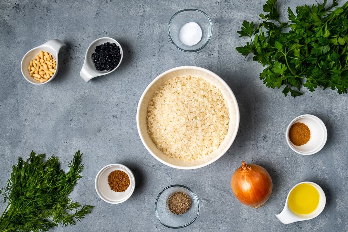 Overhead view of ingredients for Turkish iç pilav, including rice, onion, pine nuts, currants, spices, olive oil, and fresh herbs.