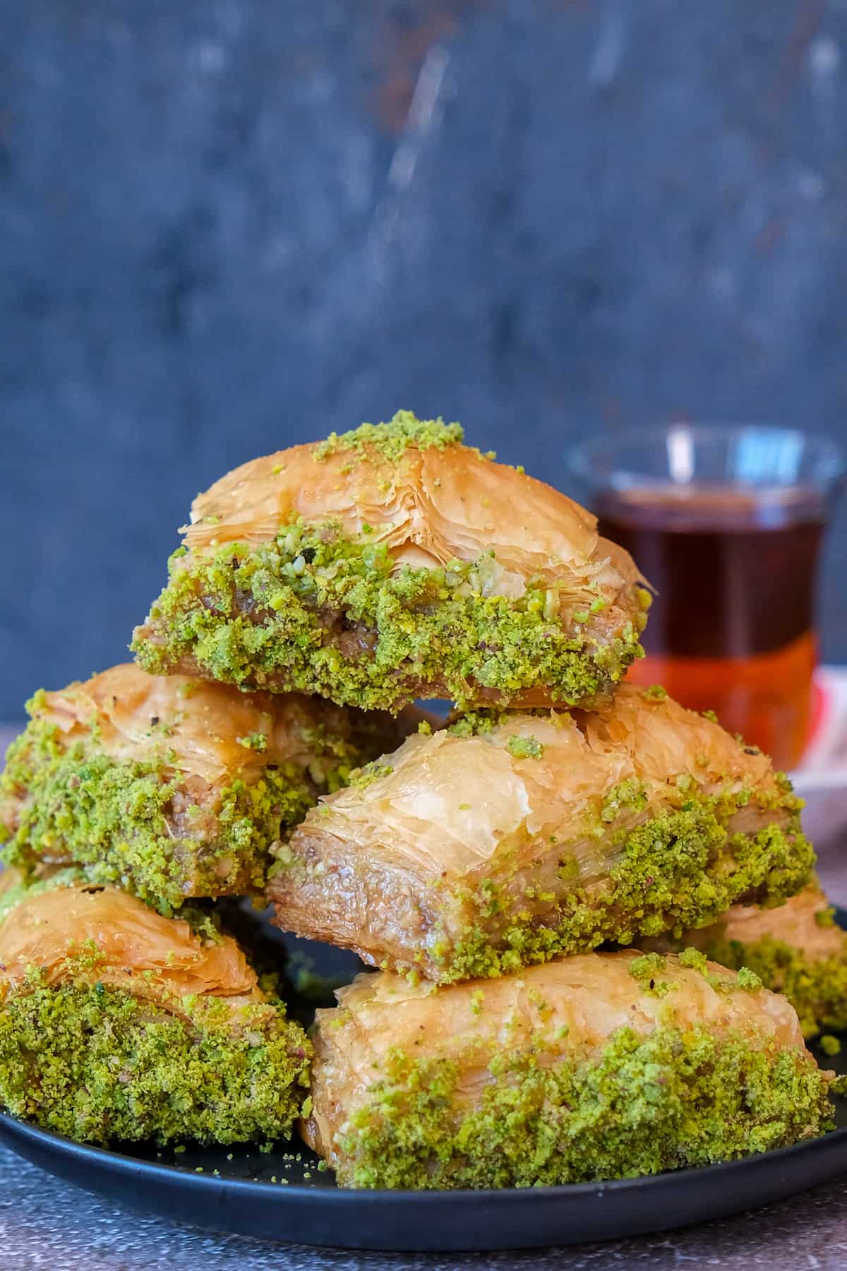 Turkish baklava with ground pistachios piled on a plate and a glass of Turkish tea behind these.