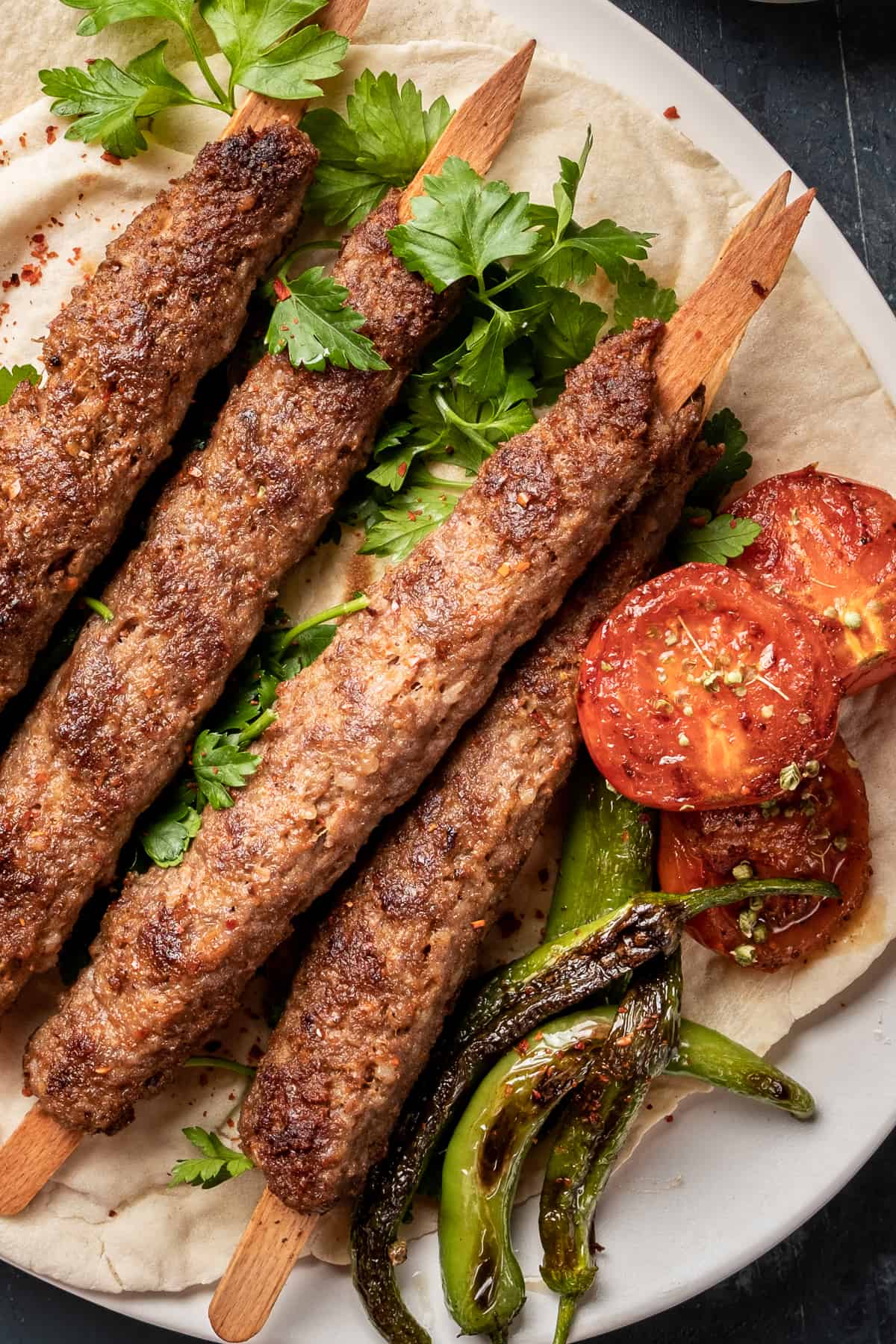Adana kebabs on wooden skewers served on a flatbread, garnished with grilled tomatoes and peppers.