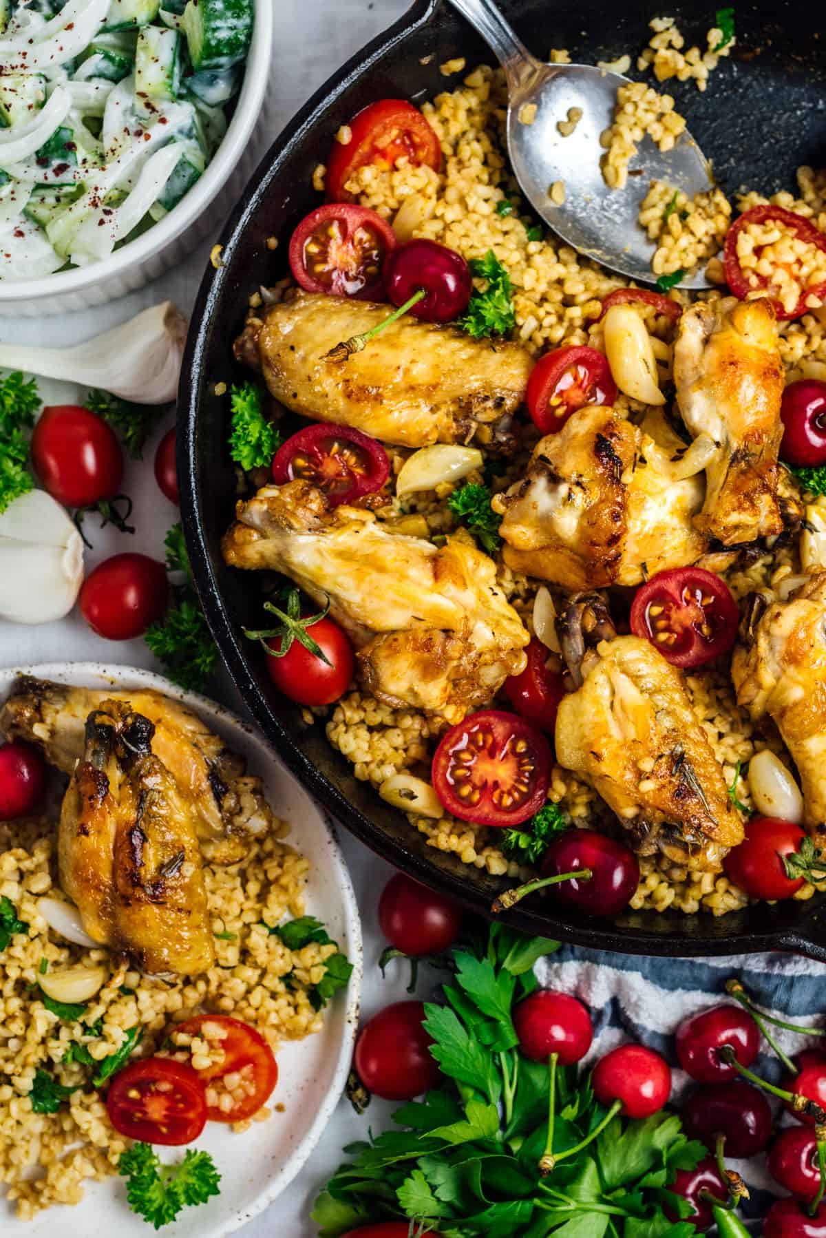 Chicken wings and bulgur wheat in a cast iron pan garnished with cherry tomatoes and parsley and a plate with this meal and another bowl with a cucumber salad on the side.