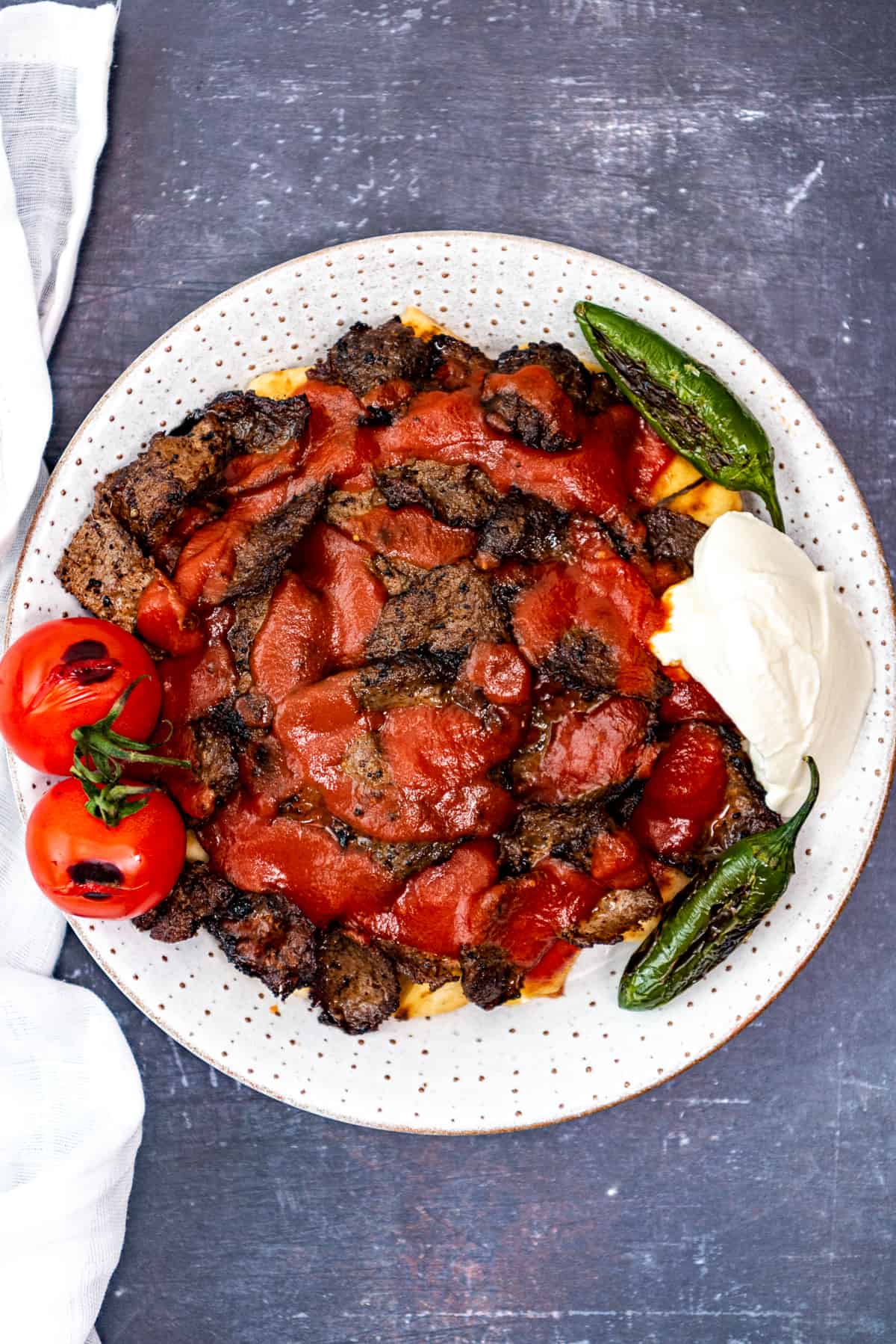 A plate loaded with iskender kebab topped with a tomato sauce and paired with yogurt and green peppers on the side.