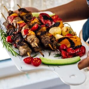 Woman holding a serving board loaded with lamb shish kebabs and grilled vegetables.