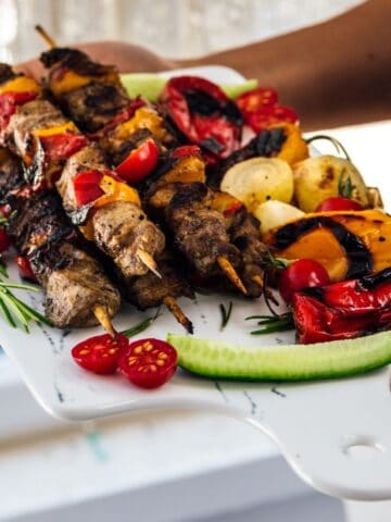 Woman holding a serving board loaded with lamb shish kebabs and grilled vegetables.