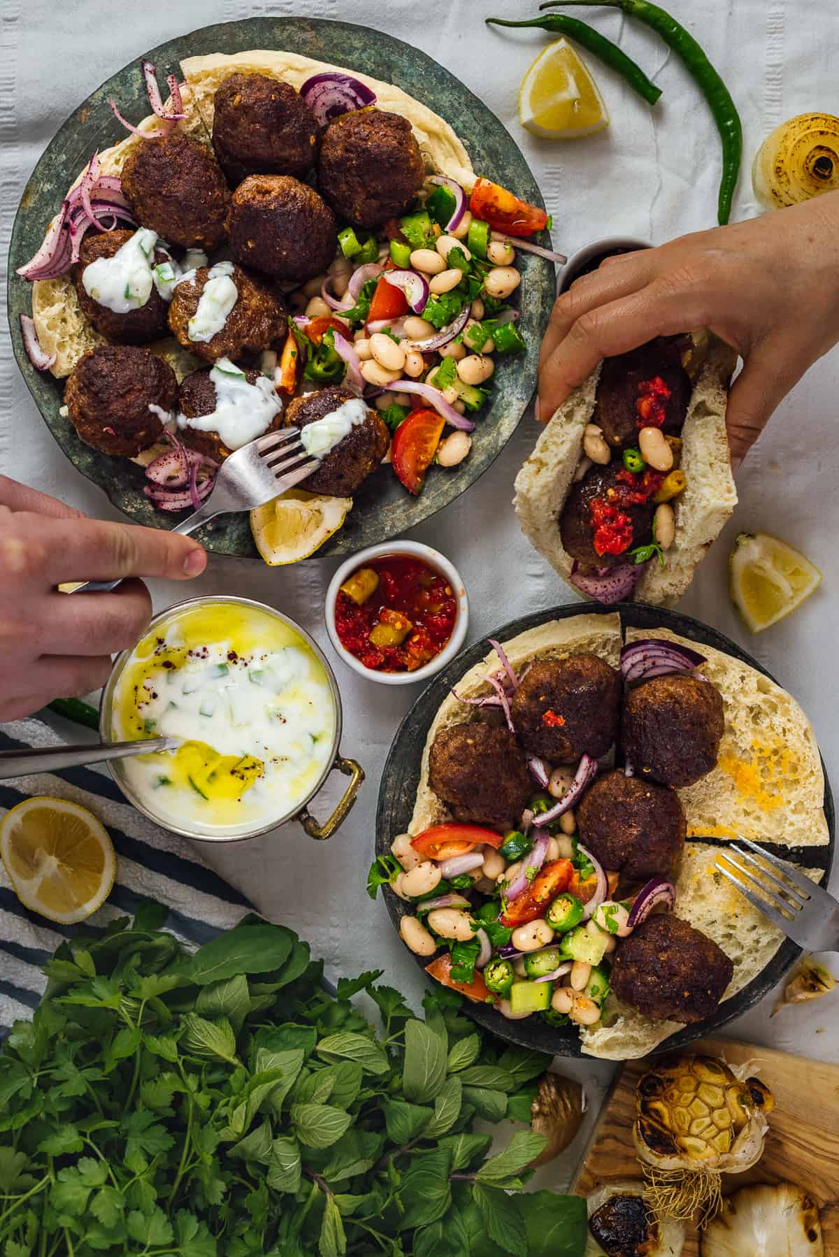 Hands taking Turkish meatballs from two plates accompanied by herbs, bean salad, yogurt dip and flat breads.