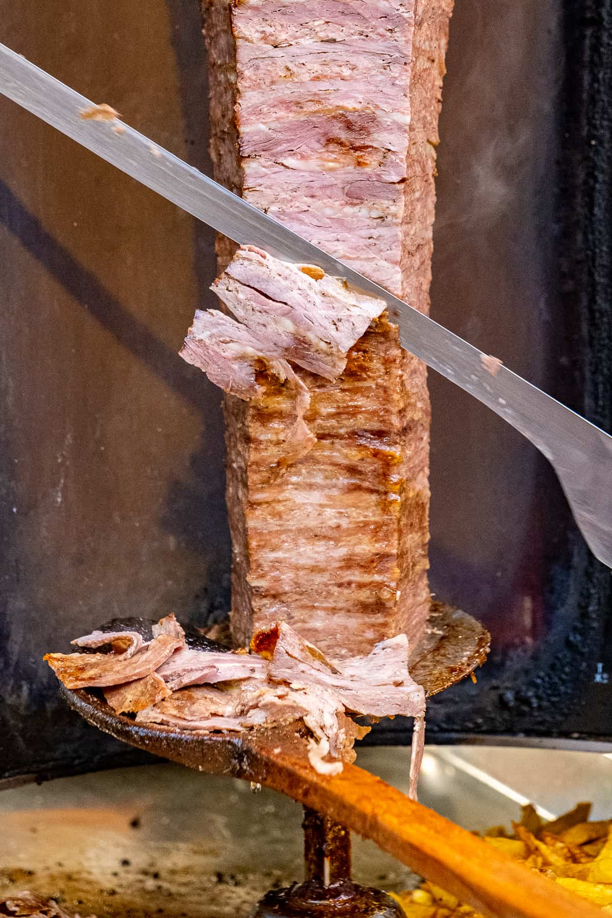 Doner kebab is being sliced with a huge knife and thin slices falling into a wooden spoon.