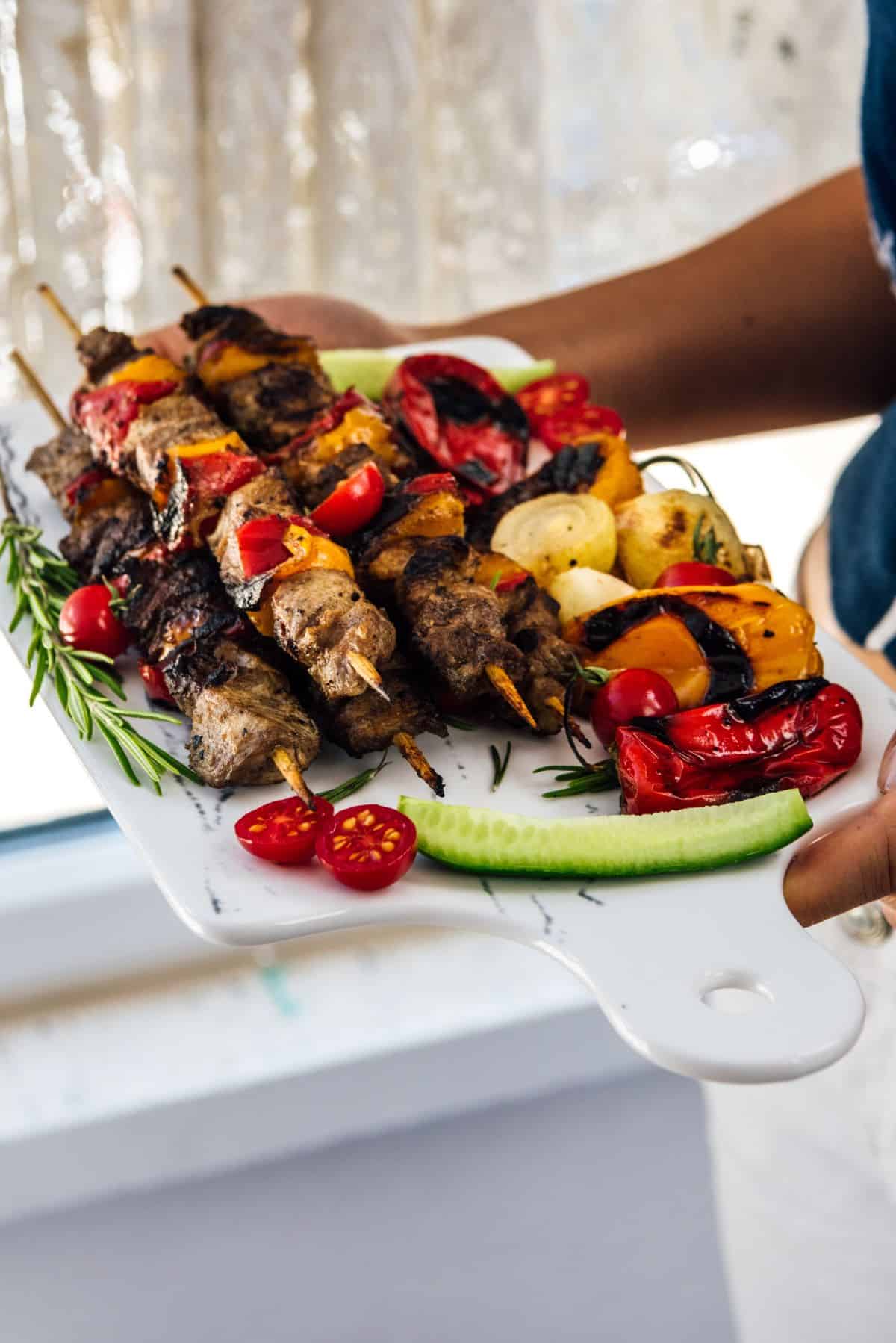 Woman holding a board loaded with lamb kebabs and roasted red and orange peppers, roasted onions, cherry tomatoes and cucumber slices.