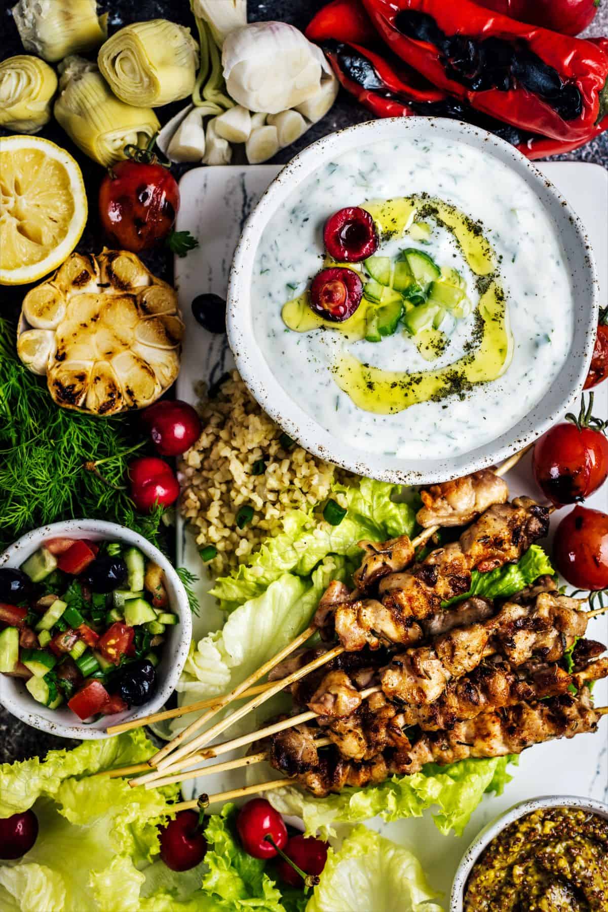 Chicken kebabs accompanied with herbs, bulgur pilaf, shepherd salad, roasted garlic, artichoke hearts and a big bowl of Turkish yogurt dip cacik.