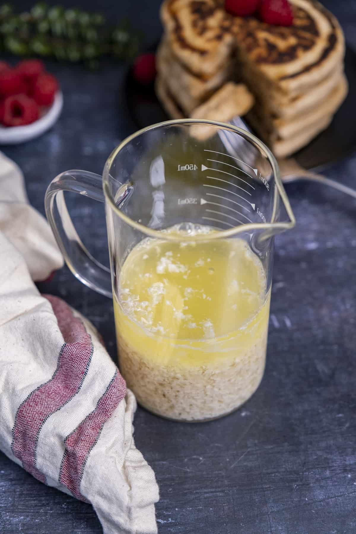 Almond buttermilk in a glass cup, a beige linen with red stripes on the side, raspberries and pancakes behind these.