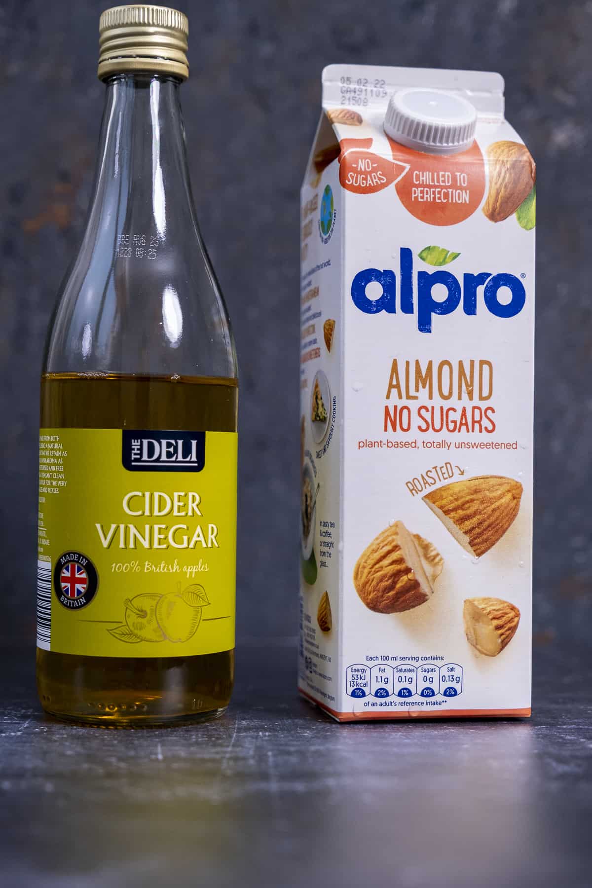 A carton of Alpro almond milk and a bottle of apple cider vinegar on a dark background.