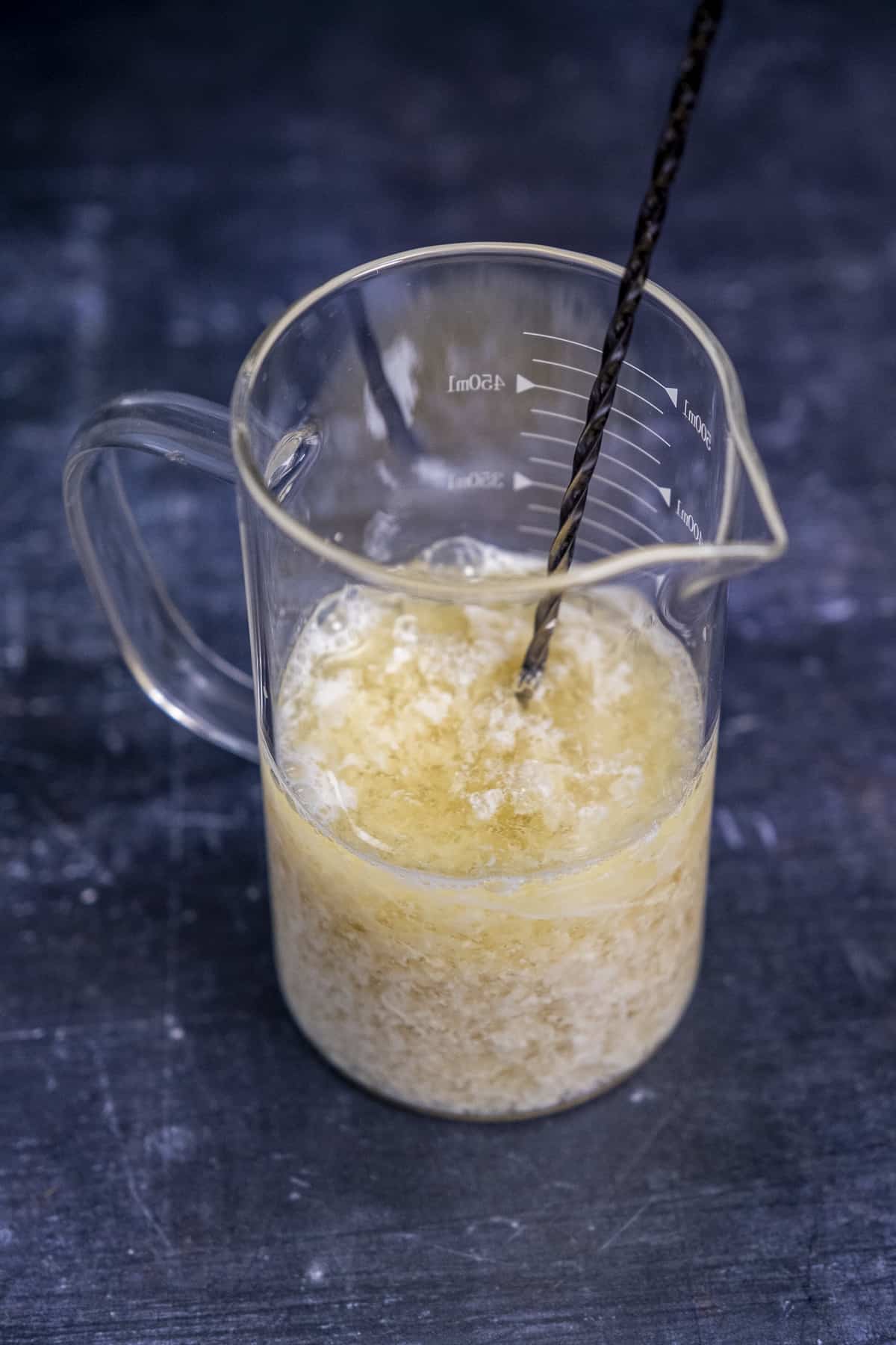 Almond milk and vinegar mixture in a glass measuring cup and a stirring spoon inside it.