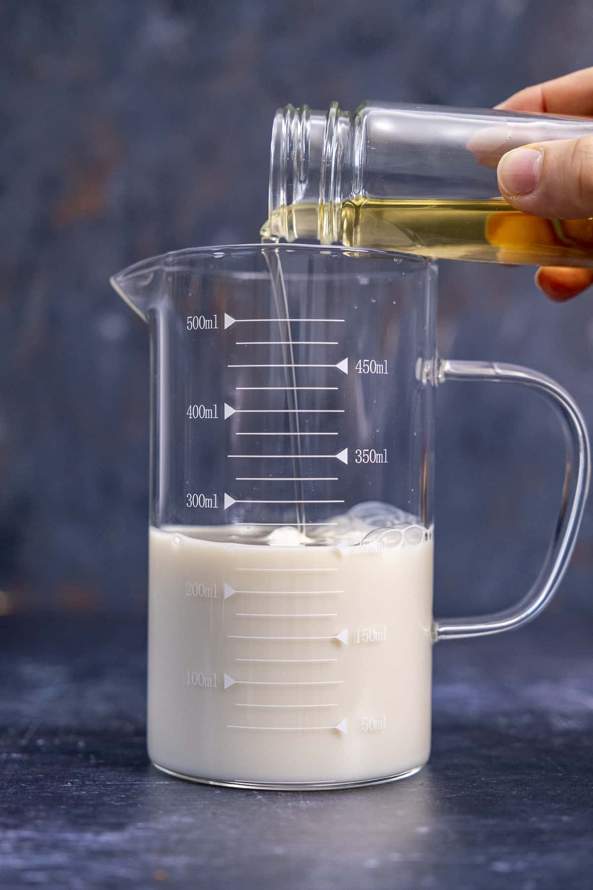 A hand pouring vinegar into almond milk in a glass measuring cup.